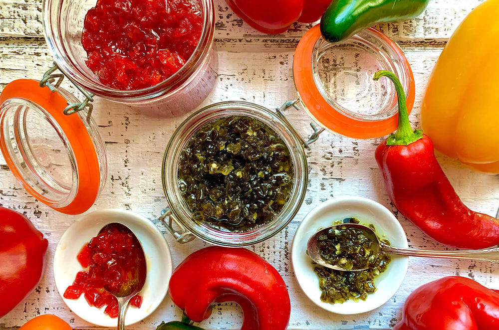Two styles of chili pepper jam—red and green—are placed in jars on a table with garden-fresh peppers surrounding them.