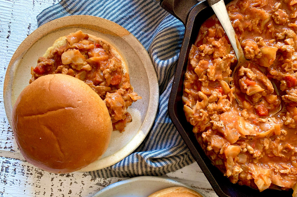 A ceramic plate with a sloppy Joe sandwich next to a skillet filled with sloppy Joe meat.